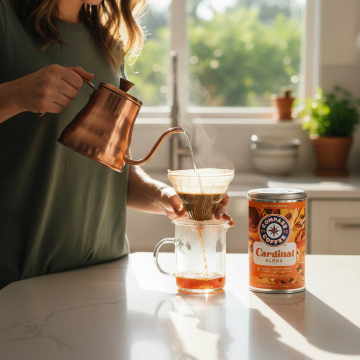 Person pouring coffee from a copper kettle into a glass mug with a can of Coffee People Cardinal Blend on a kitchen counter.